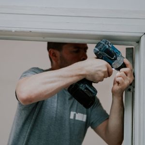 One handsome young male builder stands in a doorway and unscrews the screws of the sling with a drill, close-up side view. Concept of construction work.